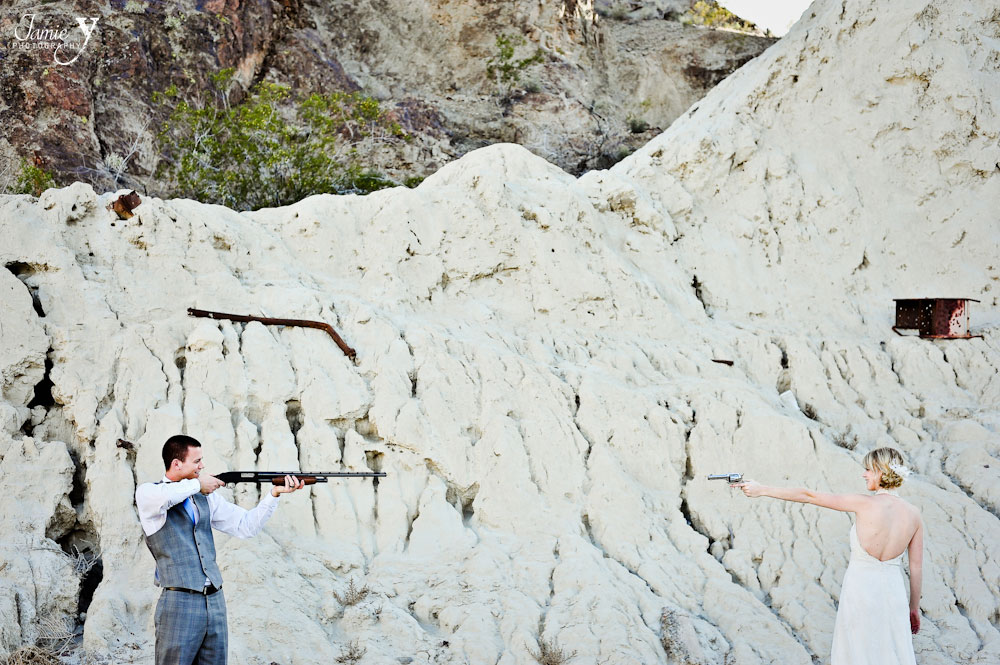 awesome photograph of bride and groom pointing guns at each other in the middle of the desert