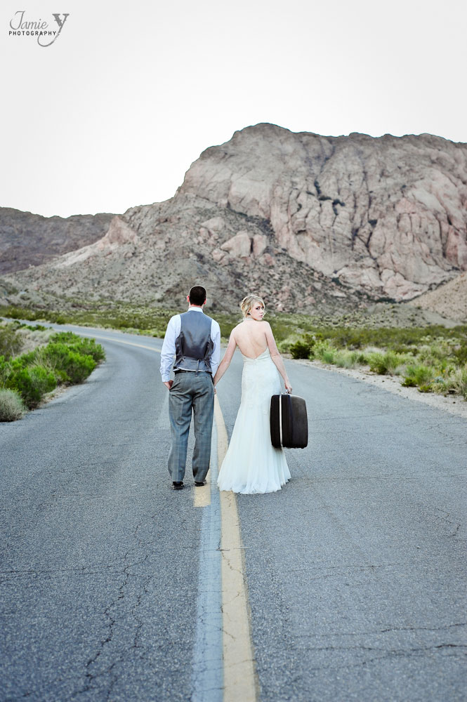 Photo of bride and groom walking down deserted street with vintage suitcase in nevada desert