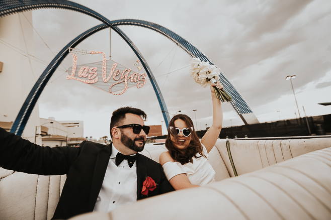 bride and groom in the back of a convertable in las vegas with hands in the air and a las vegas neon sign behind them